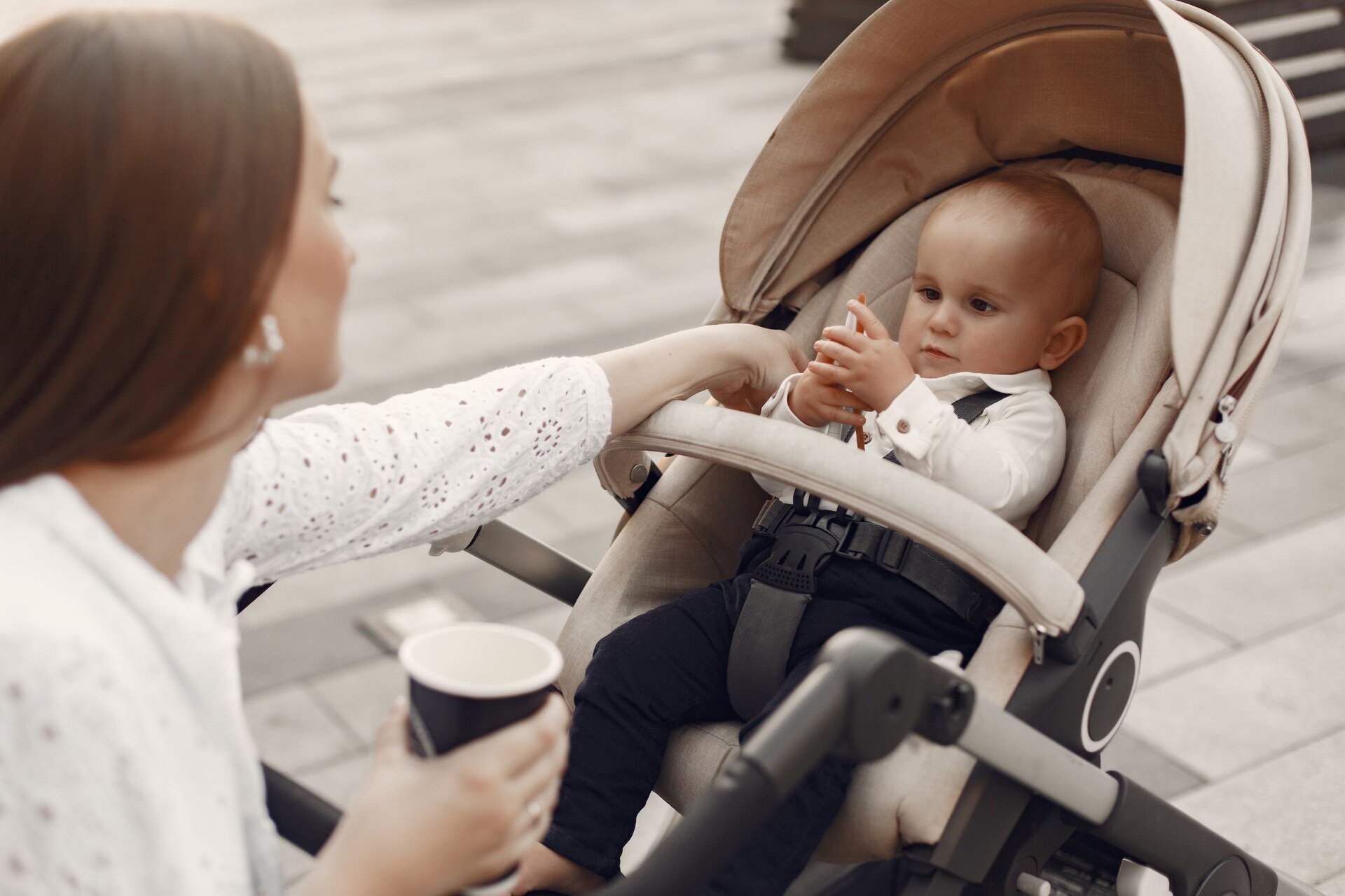 Ett litet barn sitter i en beige sittvagn med uppfälld sufflett medan mamman dricker kaffe