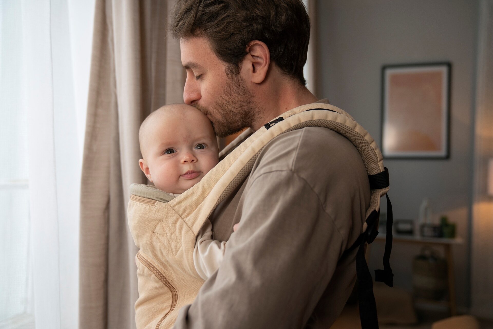 En pappa pussar sin bebis som sitter i en ljus bärsele vänd mot honom