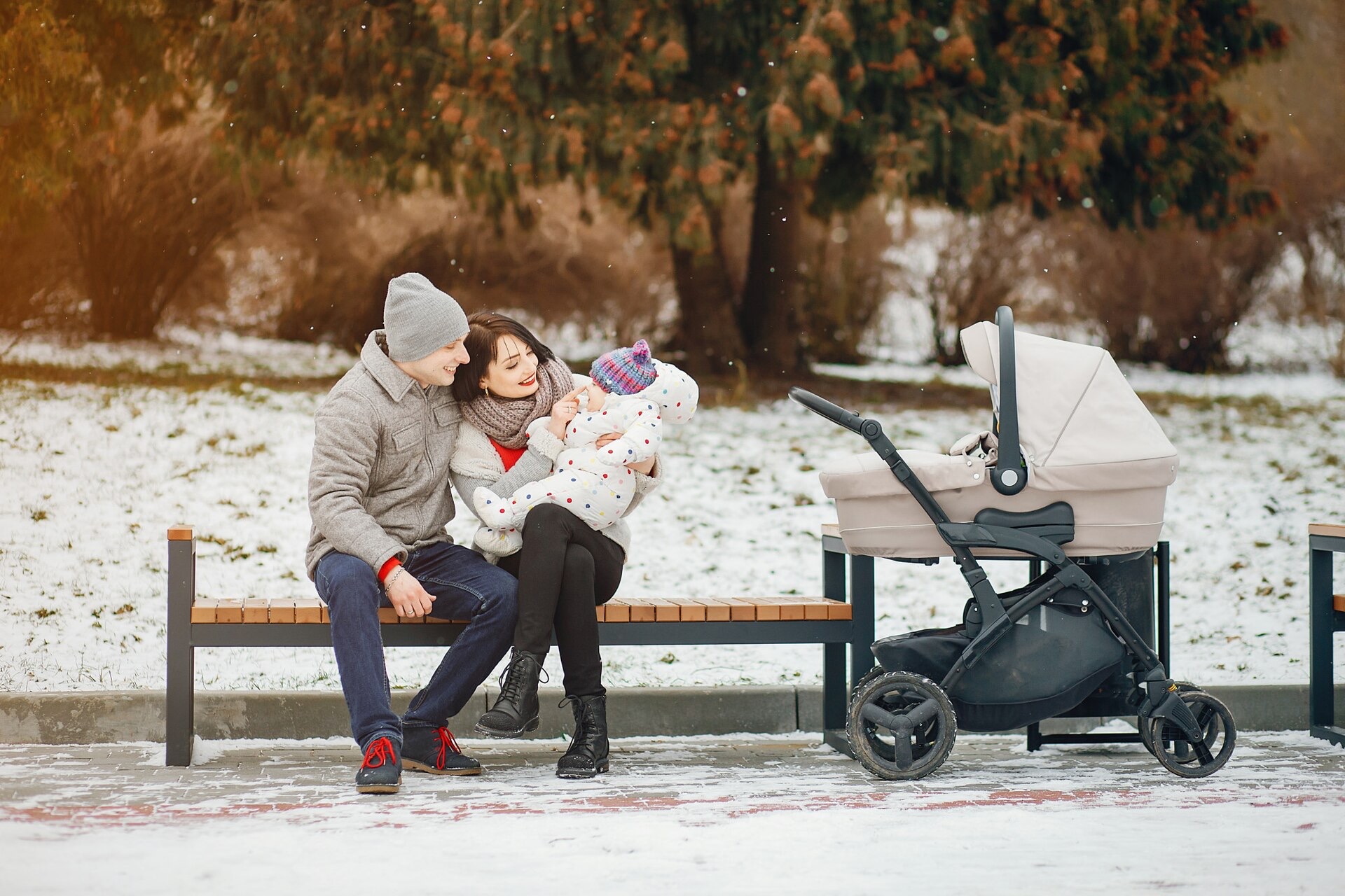 En familj sitter på en parkbänk i snön bredvid en ljus barnvagn med liggdel