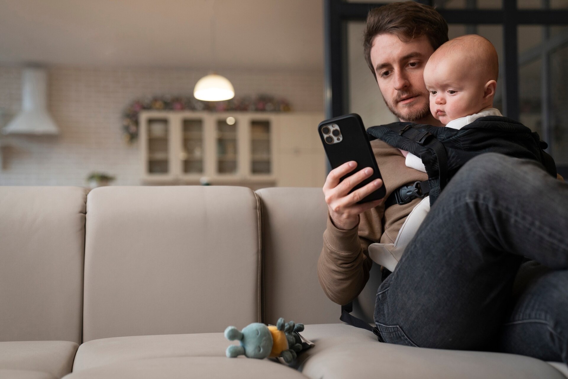 En pappa med sin bebis i en bärsele sitter i soffan och tittar på mobilen